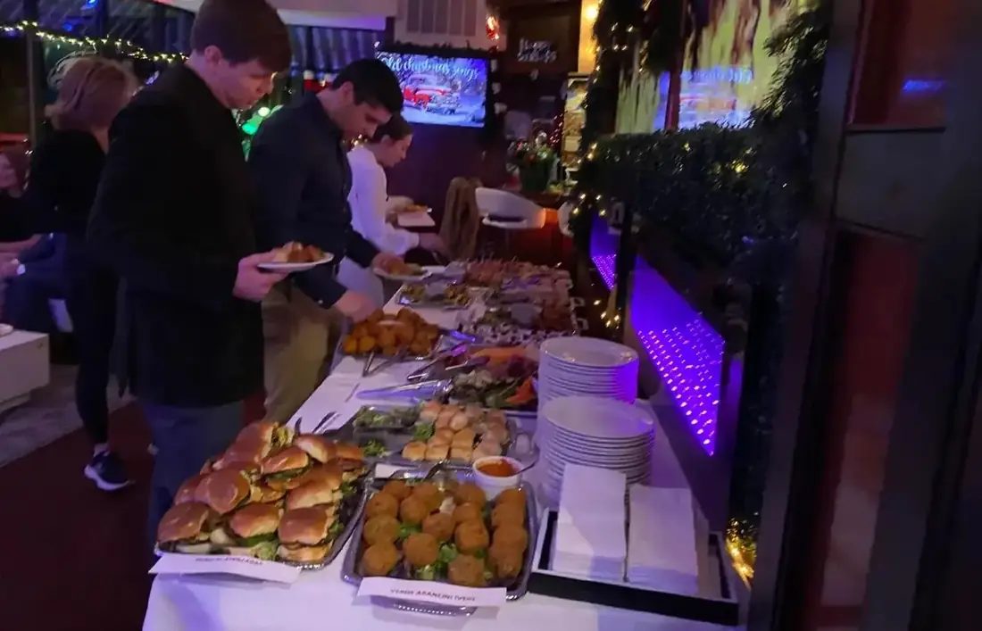 A labelled catering buffet at a corporate party — short rib sliders, verde arancini marked vegetarian and passed skewers along a white-linen table.