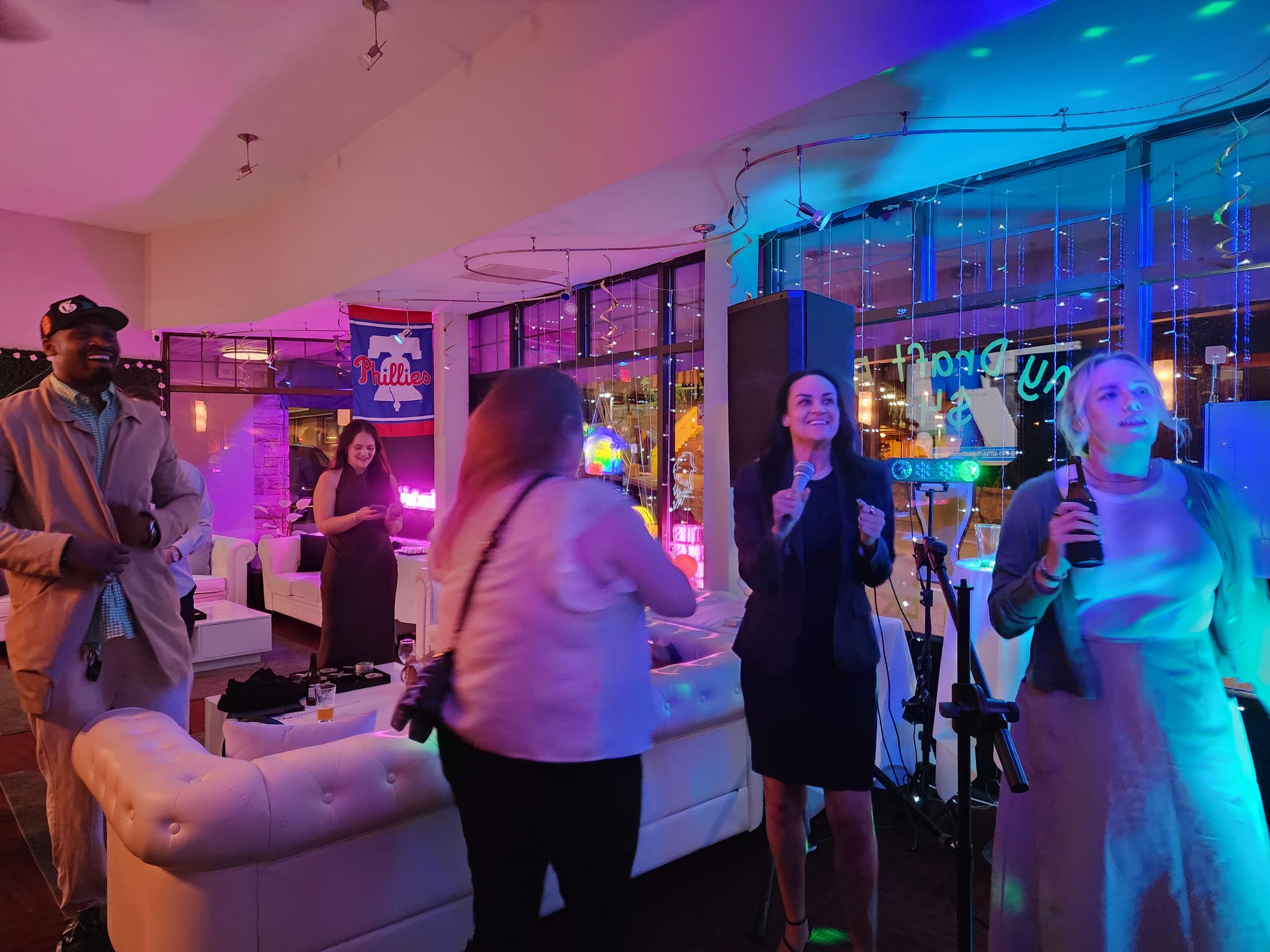 Karaoke night in the lounge at 660 LIVE — guests singing on the microphone beside white tufted couches, neon and string lights in the windows