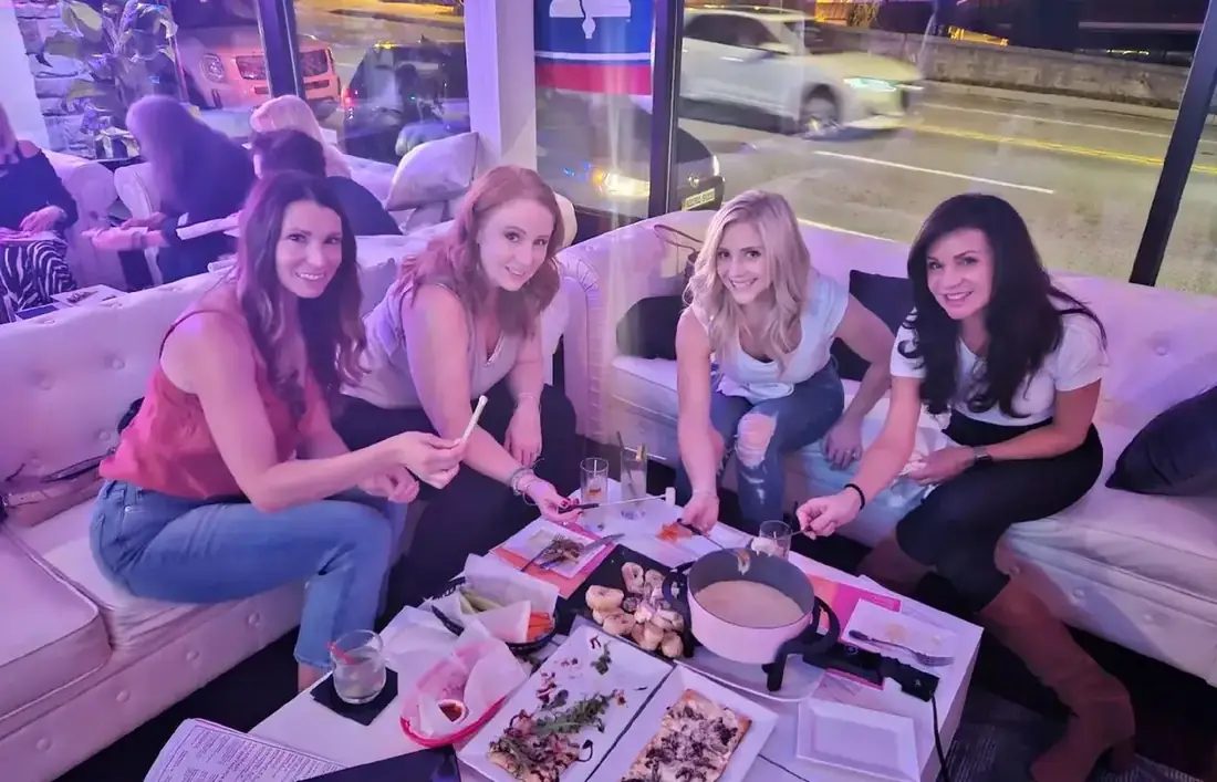 Four friends sharing fondue and flatbreads on a white sofa at 660 LIVE, Lancaster Avenue through the window behind.