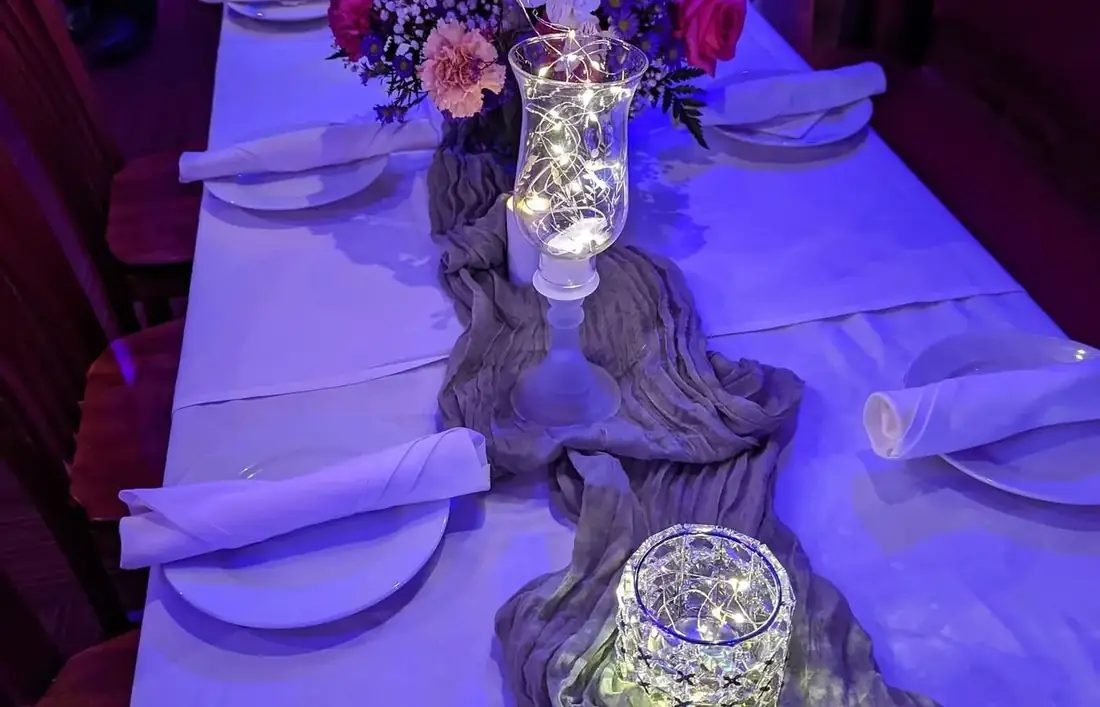 A long table styled with white linen, a cheesecloth runner, fairy lights in glass hurricanes and a floral centrepiece.